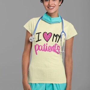 Puede incluir: Una mujer con una camiseta amarilla clara con el texto "Amo a mis pacientes" en negro y rosa. El coraz&oacute;n es rosa con un dise&ntilde;o de estampado de leopardo. Lleva un estetoscopio azul alrededor del cuello y una mascarilla quir&uacute;rgica azul claro.