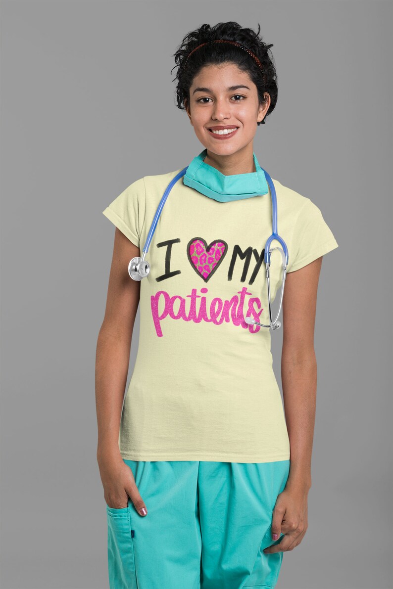 Puede incluir: Una mujer con una camiseta amarilla clara con el texto "Amo a mis pacientes" en negro y rosa. El coraz&oacute;n es rosa con un dise&ntilde;o de estampado de leopardo. Lleva un estetoscopio azul alrededor del cuello y una mascarilla quir&uacute;rgica azul claro.