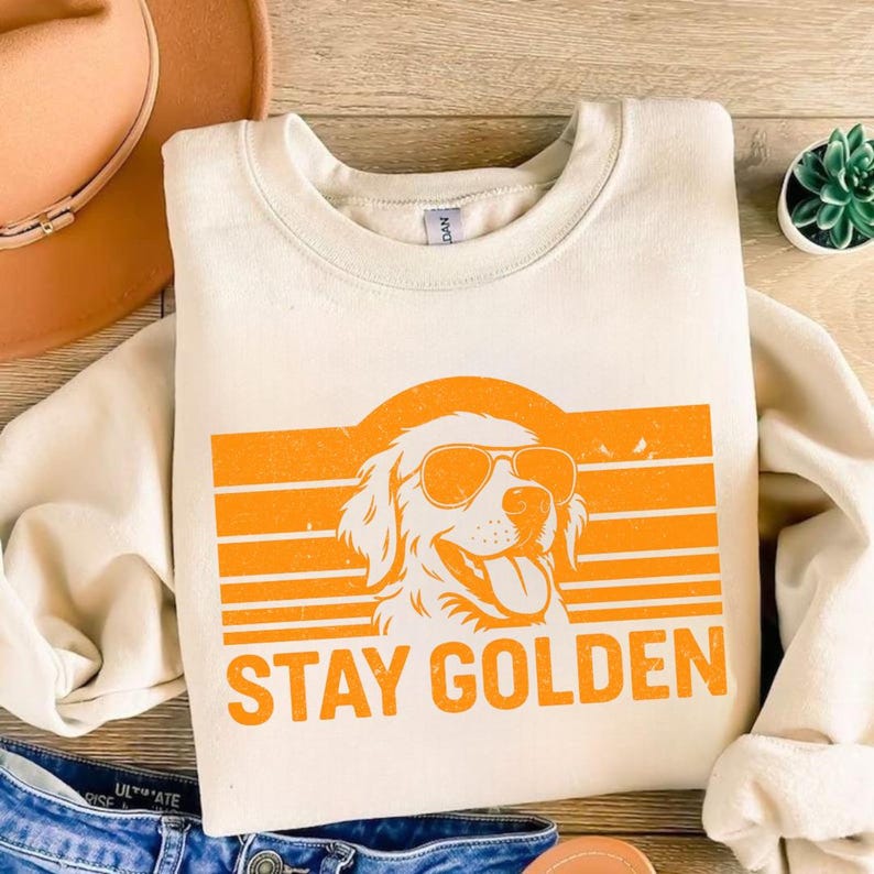 May include: Cream-colored sweatshirt with an orange graphic of a Golden Retriever wearing sunglasses, set against horizontal orange stripes. The words "STAY GOLDEN" are printed below the dog image. The sweatshirt is folded on a wooden surface.