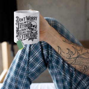 May include: White ceramic mug with the text "Don't Worry My Tattoos Don't Like You Either" in a black gothic font, with a skull and rose design. A green tea bag is attached. The mug is held by a person wearing blue plaid pajamas.