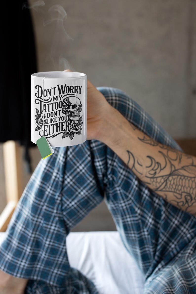 May include: White ceramic mug with the text "Don't Worry My Tattoos Don't Like You Either" in a black gothic font, with a skull and rose design. A green tea bag is attached. The mug is held by a person wearing blue plaid pajamas.