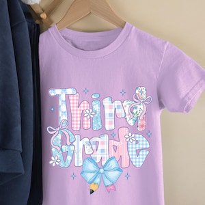Puede incluir: Una camiseta de color lila claro con la frase "Third Grade" en un dise&ntilde;o colorido y estampado. El texto presenta detalles florales, a cuadros y lazos. La camiseta est&aacute; colgada en una percha de madera.