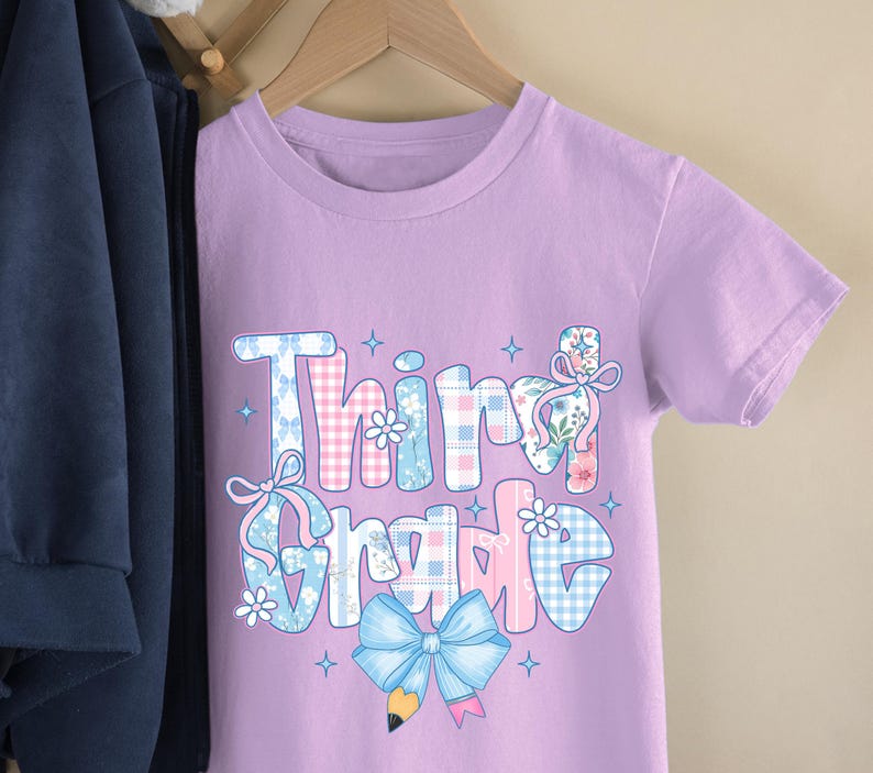 Puede incluir: Una camiseta de color lila claro con la frase "Third Grade" en un dise&ntilde;o colorido y estampado. El texto presenta detalles florales, a cuadros y lazos. La camiseta est&aacute; colgada en una percha de madera.