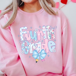May include: Pink Fourth Grade sweatshirt with a colorful graphic. The design features the words "Fourth Grade" in a playful font, decorated with bows, flowers, and a pencil. The letters have a patchwork pattern in blue, pink, and white.