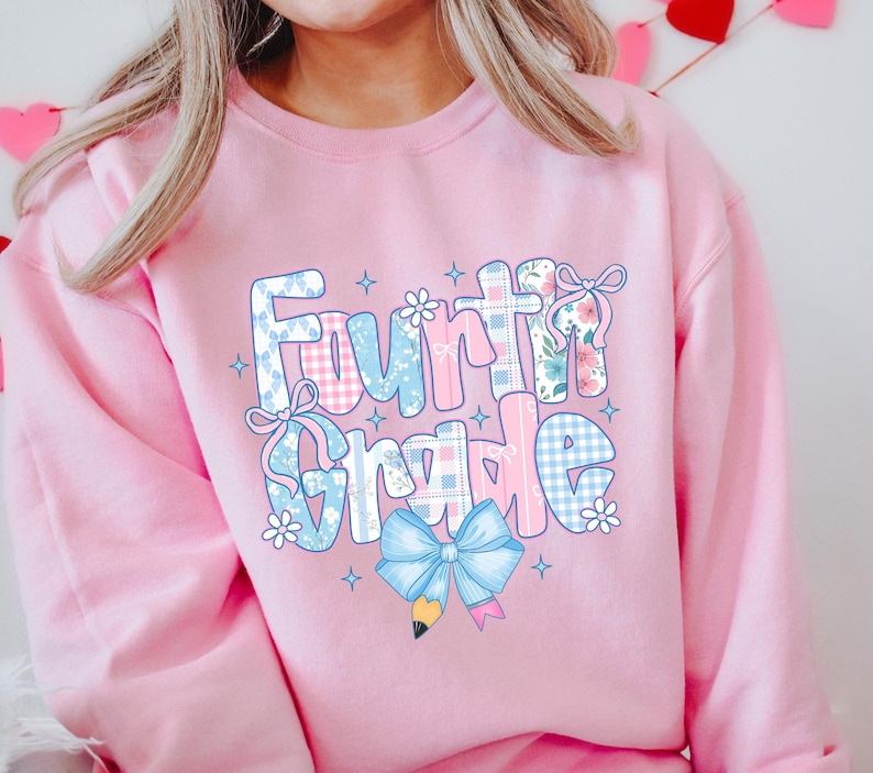 May include: Pink Fourth Grade sweatshirt with a colorful graphic. The design features the words "Fourth Grade" in a playful font, decorated with bows, flowers, and a pencil. The letters have a patchwork pattern in blue, pink, and white.