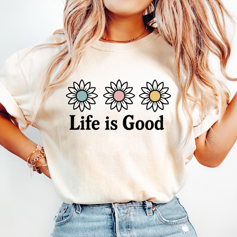 May include: A cream-coloured t-shirt displaying three daisy graphics with pastel centres and the text "Life is Good" in black. The daisies have blue, pink, and yellow centres. The shirt has rolled-up sleeves and is paired with blue jeans.