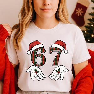 May include: White t-shirt featuring the number "67" in a red and green plaid pattern, each number topped with a Santa hat. Below the numbers are cartoon hands with holly sprigs. The shirt is a festive holiday design.