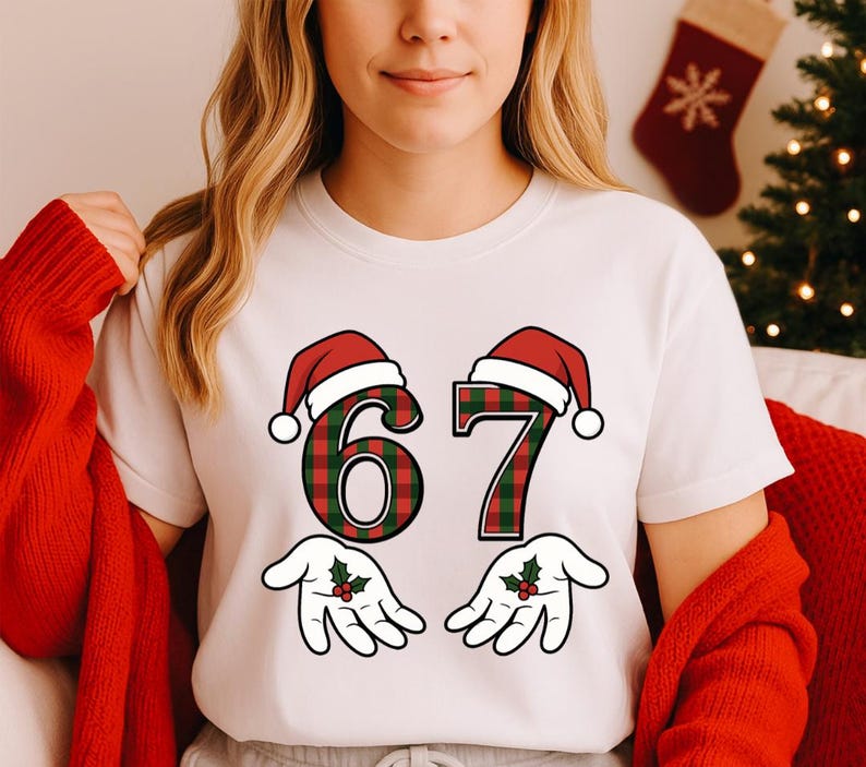 May include: White t-shirt featuring the number "67" in a red and green plaid pattern, each number topped with a Santa hat. Below the numbers are cartoon hands with holly sprigs. The shirt is a festive holiday design.