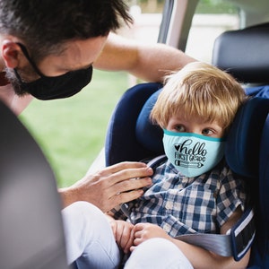May include: A young boy wearing a blue face mask with the text "hello first grade" and a pencil graphic sits in a car seat. An adult is helping to secure the child in the seat.