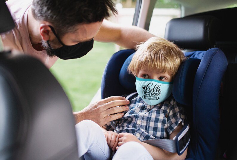May include: A young boy wearing a blue face mask with the text "hello first grade" and a pencil graphic sits in a car seat. An adult is helping to secure the child in the seat.
