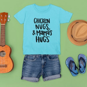 May include: A light blue t-shirt with the text "Chicken Nugs. & Mamas Hugs" in black. The shirt is laid out on a green background with a brown hat, a ukulele, and blue flip-flops.