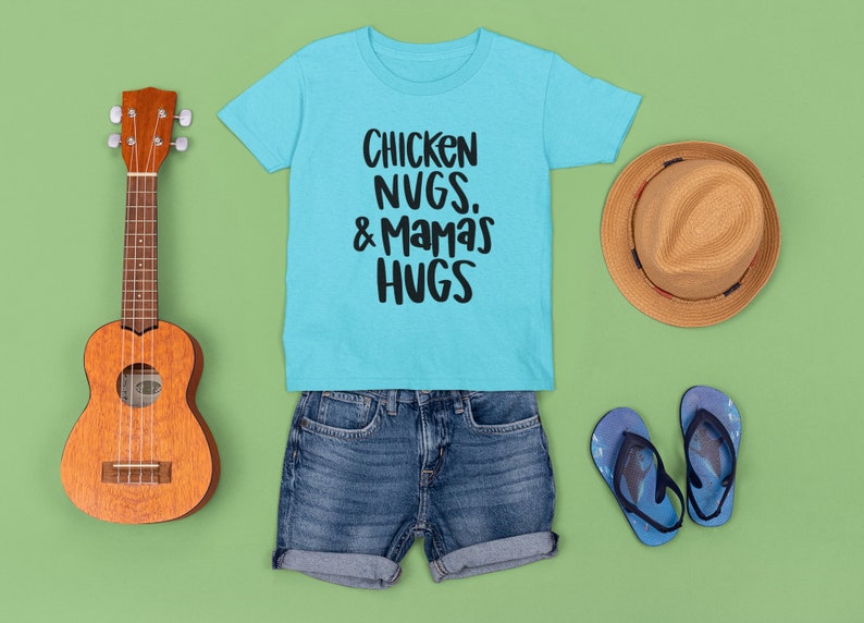 May include: A light blue t-shirt with the text "Chicken Nugs. & Mamas Hugs" in black. The shirt is laid out on a green background with a brown hat, a ukulele, and blue flip-flops.