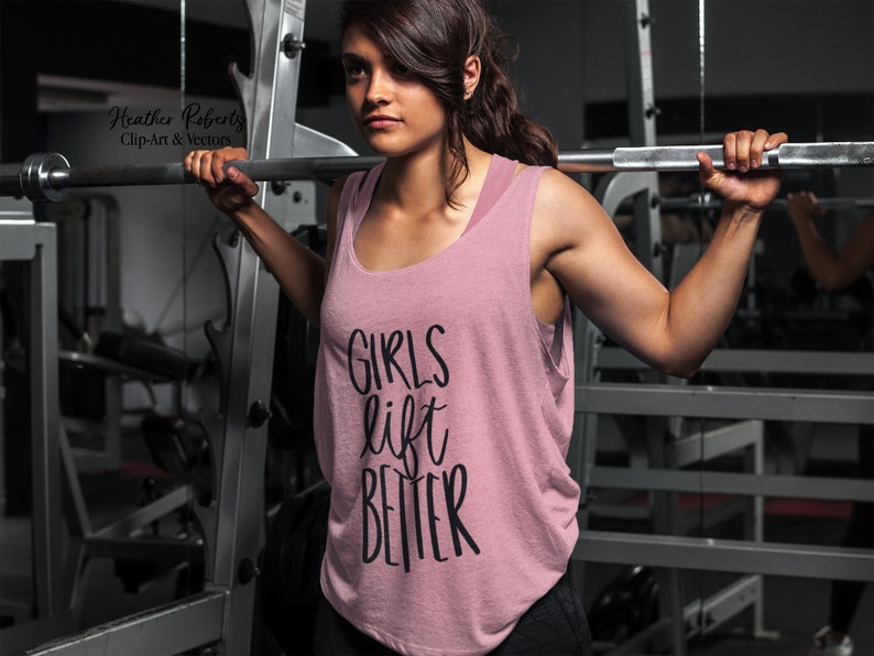 K&ouml;nnte beinhalten: Ein staubrosa Tanktop mit dem Text "GIRLS lift BETTER" in schwarzer Schrift. Das Tanktop wird in einem Fitnessstudio getragen, mit Gewichtheberger&auml;ten im Hintergrund.