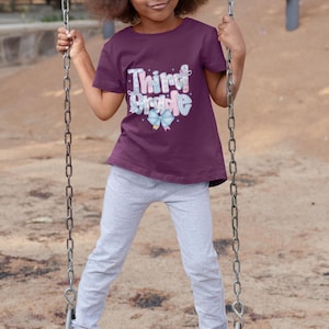 Puede incluir: Una ni&ntilde;a en un columpio, con una camiseta granate con las palabras "Third Grade" en un dise&ntilde;o colorido. Lleva pantalones de ch&aacute;ndal grises y zapatillas rosas, con un lazo rosa en el pelo.