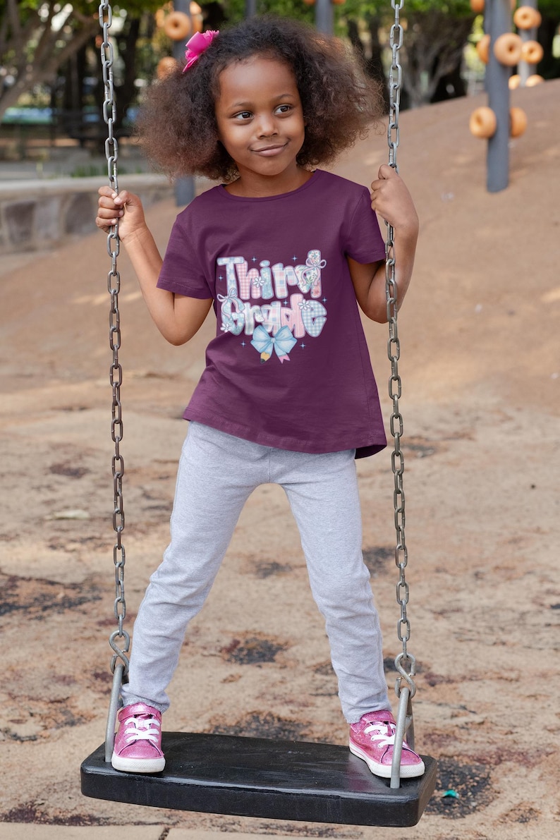 Puede incluir: Una ni&ntilde;a en un columpio, con una camiseta granate con las palabras "Third Grade" en un dise&ntilde;o colorido. Lleva pantalones de ch&aacute;ndal grises y zapatillas rosas, con un lazo rosa en el pelo.