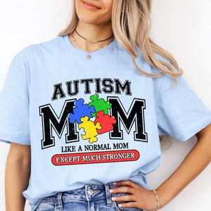 May include: Light blue t-shirt with the text "Autism Mom" in black and white with a colorful puzzle piece design. The text below reads "Like a normal mom except much stronger."