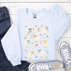 May include: A light blue sweatshirt featuring a charming pattern of white geese adorned with colorful bows and surrounded by floral accents. The crew neck sweatshirt is made of a soft material and is a comfortable addition to any wardrobe.