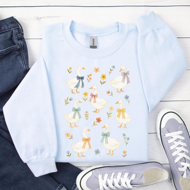 May include: A light blue sweatshirt featuring a charming pattern of white geese adorned with colorful bows and surrounded by floral accents. The crew neck sweatshirt is made of a soft material and is a comfortable addition to any wardrobe.