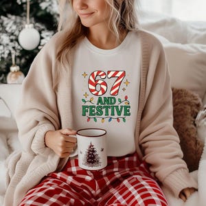 May include: A white t-shirt with the text "67 AND FESTIVE" in a candy cane and Christmas light design. The person is wearing a beige cardigan and red plaid pajama pants. A white mug with a Christmas tree design is held in the person's hand.