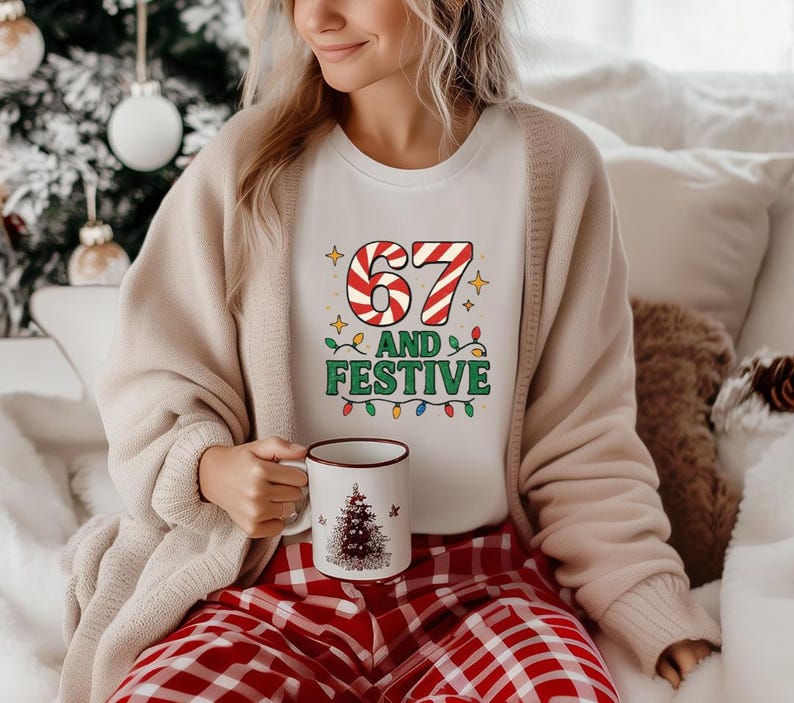 May include: A white t-shirt with the text "67 AND FESTIVE" in a candy cane and Christmas light design. The person is wearing a beige cardigan and red plaid pajama pants. A white mug with a Christmas tree design is held in the person's hand.