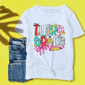 Puede incluir: Camiseta blanca con un gr&aacute;fico colorido que dice "Third Grade" con un l&aacute;piz, un lazo y otros dise&ntilde;os de tem&aacute;tica escolar.