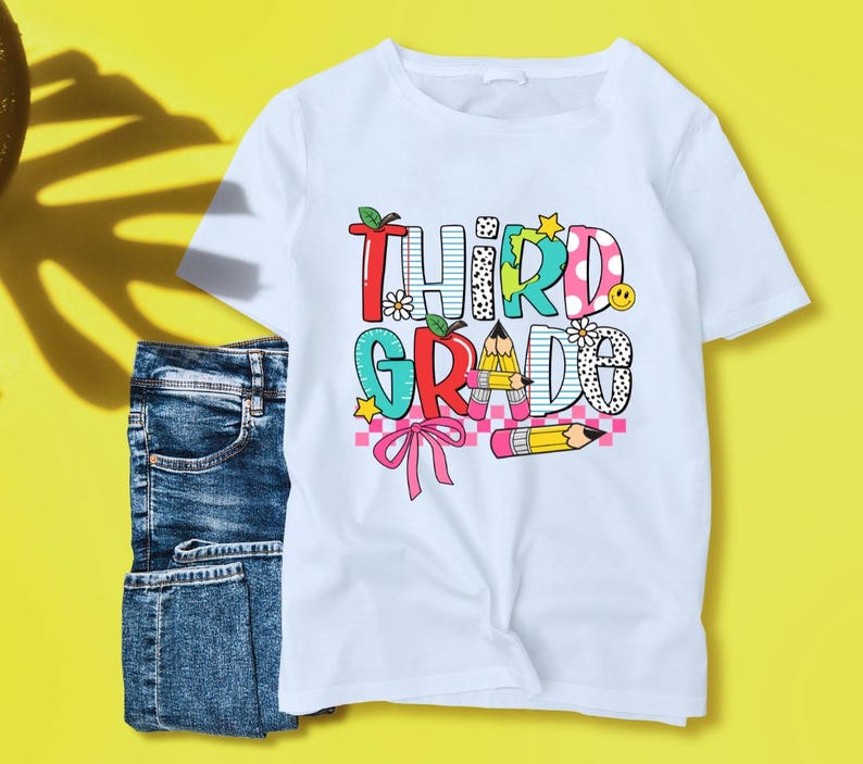 Puede incluir: Camiseta blanca con un gr&aacute;fico colorido que dice "Third Grade" con un l&aacute;piz, un lazo y otros dise&ntilde;os de tem&aacute;tica escolar.