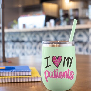 Puede incluir: Un vaso de vino de acero inoxidable verde claro con un dise&ntilde;o de coraz&oacute;n negro y rosa y el texto "Amo a mis pacientes". El vaso tiene una pajita y est&aacute; sobre una mesa de madera.