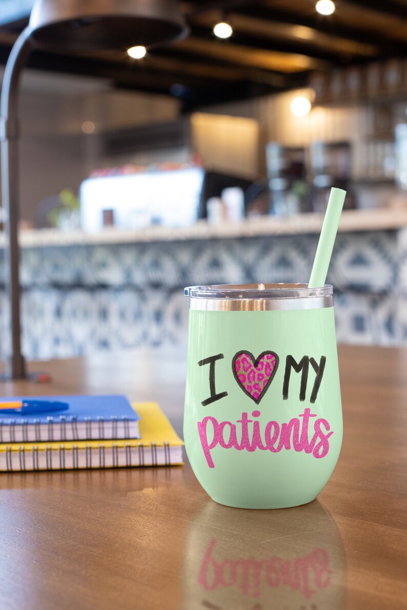 Puede incluir: Un vaso de vino de acero inoxidable verde claro con un dise&ntilde;o de coraz&oacute;n negro y rosa y el texto "Amo a mis pacientes". El vaso tiene una pajita y est&aacute; sobre una mesa de madera.