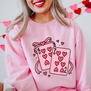 May include: Pink sweatshirt featuring a graphic of two playing cards with heart symbols. The cards are tied together with a pink ribbon and bow. The number 6 and 7 are visible on the cards. Small hearts are scattered around the cards.