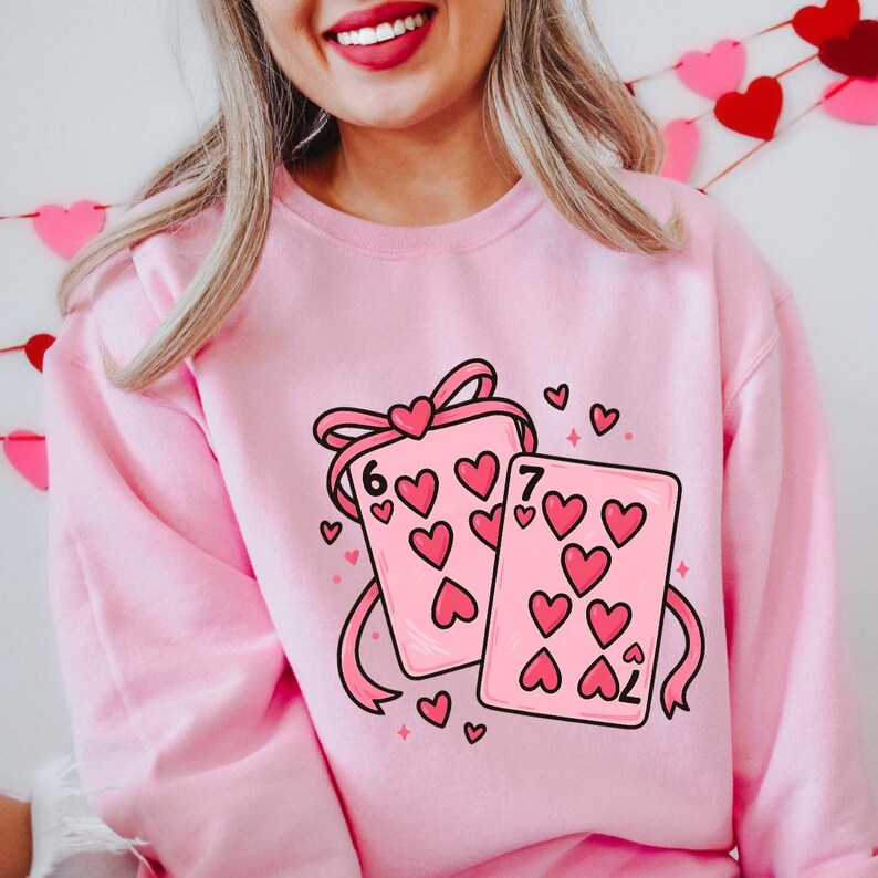 May include: Pink sweatshirt featuring a graphic of two playing cards with heart symbols. The cards are tied together with a pink ribbon and bow. The number 6 and 7 are visible on the cards. Small hearts are scattered around the cards.