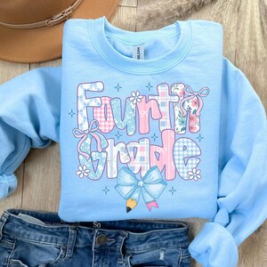 May include: A light blue Fourth Grade sweatshirt with a colorful graphic. The graphic features the words "Fourth Grade" in a mix of patterns, including plaid and floral designs, with bows and a pencil illustration.