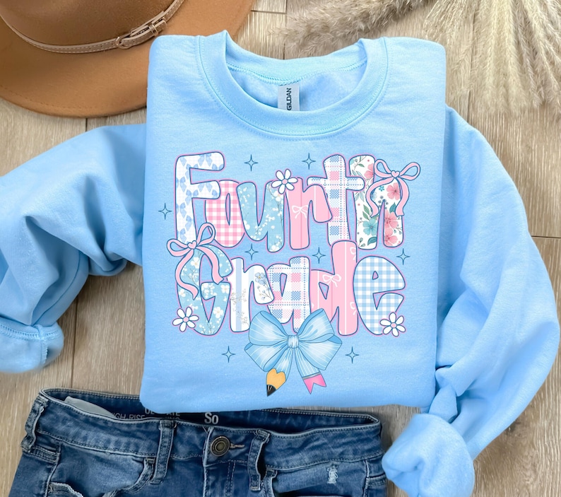 May include: A light blue Fourth Grade sweatshirt with a colorful graphic. The graphic features the words "Fourth Grade" in a mix of patterns, including plaid and floral designs, with bows and a pencil illustration.
