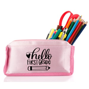 May include: Pink pencil case with a zipper closure. The case has a black graphic that says "hello first grade" with a heart and a pencil. The case is filled with colored pencils, markers, and a pair of scissors.