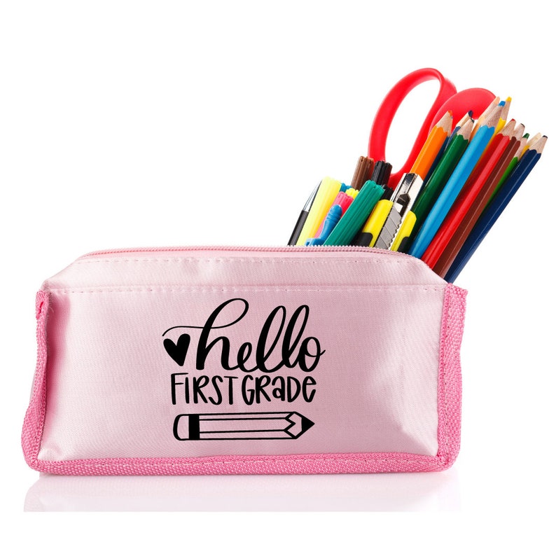 May include: Pink pencil case with a zipper closure. The case has a black graphic that says "hello first grade" with a heart and a pencil. The case is filled with colored pencils, markers, and a pair of scissors.