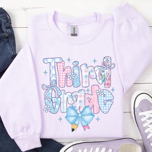 Puede incluir: Una sudadera lila claro con la frase "Third Grade" en un dise&ntilde;o de patchwork. Las letras est&aacute;n decoradas con estampados florales, lazos y cuadros vichy en tonos rosas y azules. Un lazo azul claro y un l&aacute;piz gr&aacute;fico en la parte inferior.