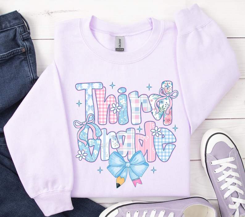 Puede incluir: Una sudadera lila claro con la frase "Third Grade" en un dise&ntilde;o de patchwork. Las letras est&aacute;n decoradas con estampados florales, lazos y cuadros vichy en tonos rosas y azules. Un lazo azul claro y un l&aacute;piz gr&aacute;fico en la parte inferior.
