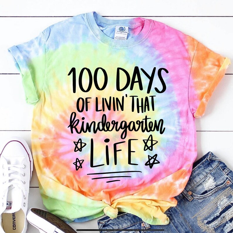 May include: A tie-dye t-shirt with the text "100 Days of Livin' That Kindergarten Life" in black lettering. The shirt has a rainbow tie-dye pattern with shades of pink, yellow, blue, and green.