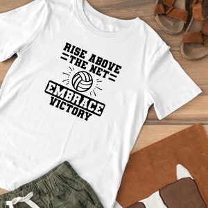 K&ouml;nnte beinhalten: Wei&szlig;es T-Shirt mit schwarzem Text, der "Rise Above The Net = Embrace Victory" lautet. Eine schwarz-wei&szlig;e Volleyballgrafik befindet sich mittig &uuml;ber dem Text.