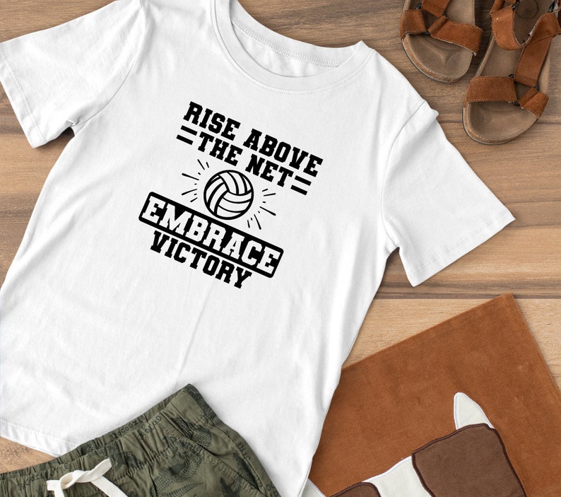 K&ouml;nnte beinhalten: Wei&szlig;es T-Shirt mit schwarzem Text, der "Rise Above The Net = Embrace Victory" lautet. Eine schwarz-wei&szlig;e Volleyballgrafik befindet sich mittig &uuml;ber dem Text.
