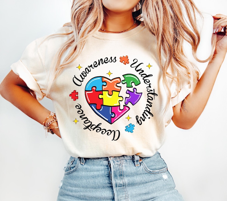 Pode incluir: Uma camiseta branca com um design de cora&ccedil;&atilde;o de pe&ccedil;as de quebra-cabe&ccedil;a coloridas. O texto ao redor do cora&ccedil;&atilde;o diz "Awareness, Understanding, Acceptance".