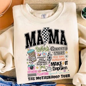 May include: A cream-colored sweatshirt with a graphic print that says "MAMA Work Grocery Store Field Trips Playground Make It Happen Kitchen Practice Sport Games The Motherhood Tour". The graphic is in a retro style with colorful accents and includes a cartoon coffee cup, a camera, a donut, and a heart.