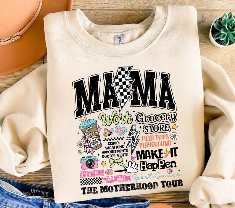 May include: A cream-colored sweatshirt with a graphic print that says "MAMA Work Grocery Store Field Trips Playground Make It Happen Kitchen Practice Sport Games The Motherhood Tour". The graphic is in a retro style with colorful accents and includes a cartoon coffee cup, a camera, a donut, and a heart.