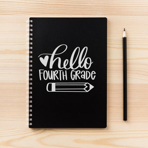 以下が含まれることがあります： 黒いスパイラルノートに「hello fourth grade」という文字が白く書かれています。ノートの隣には木製の表面に鉛筆が置かれています。