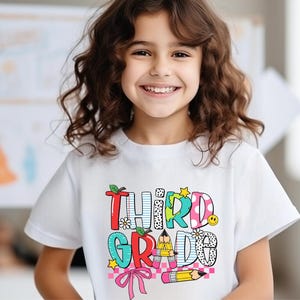 Puede incluir: Una camiseta blanca con un gr&aacute;fico colorido que dice "Tercer Grado" con un l&aacute;piz, una flor y una estrella. La camiseta la lleva puesta una ni&ntilde;a con el pelo rizado casta&ntilde;o.