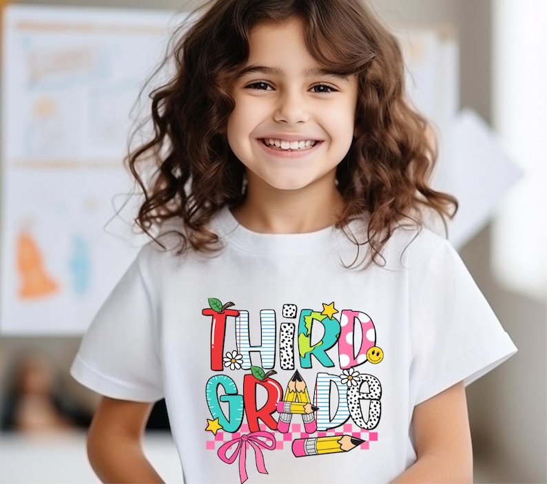 Puede incluir: Una camiseta blanca con un gr&aacute;fico colorido que dice "Tercer Grado" con un l&aacute;piz, una flor y una estrella. La camiseta la lleva puesta una ni&ntilde;a con el pelo rizado casta&ntilde;o.