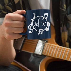 May include: A blue ceramic mug with a white design of musical notes and the letters "AFC" in a circle. The mug is being held by a person wearing a floral shirt.