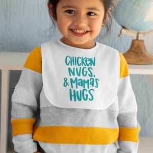 May include: A white baby bib with the text "Chicken Nugs. & Mama's Hugs" in teal blue lettering. A young child is wearing the bib and smiling.
