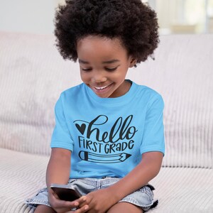 May include: A young child wearing a light blue t-shirt with the text "hello first grade" and a black heart and pencil graphic. The child is sitting on a couch and looking down at a cell phone.