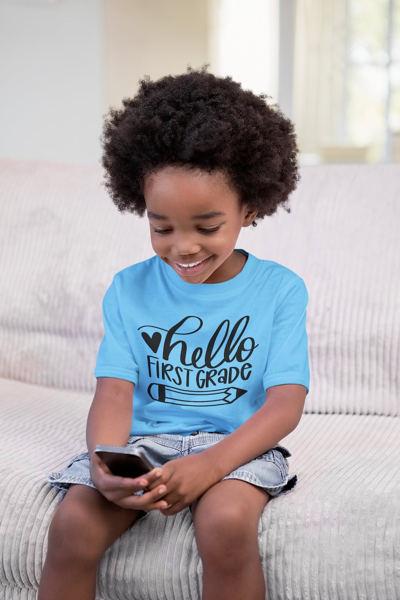 May include: A young child wearing a light blue t-shirt with the text "hello first grade" and a black heart and pencil graphic. The child is sitting on a couch and looking down at a cell phone.