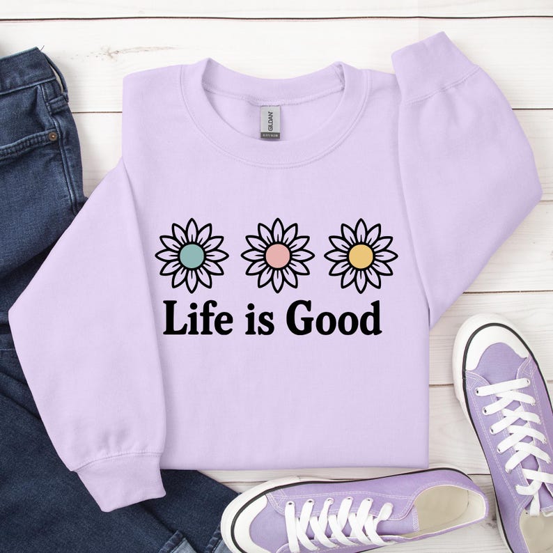 May include: A lilac sweatshirt featuring the text "Life is Good" in black, with three daisy motifs above. The flowers have pastel-coloured centres. A pair of purple trainers and blue jeans are next to the sweatshirt.